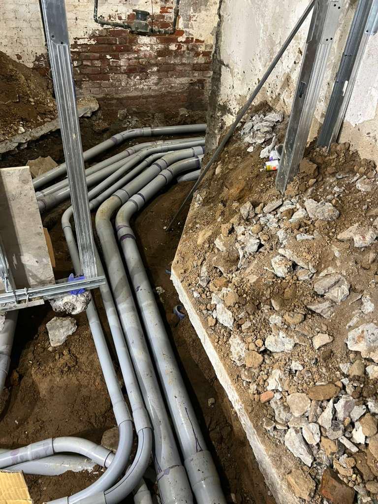 Banks of grey electrical conduit sweeping through an excavated cellar trench beside metal stud framing